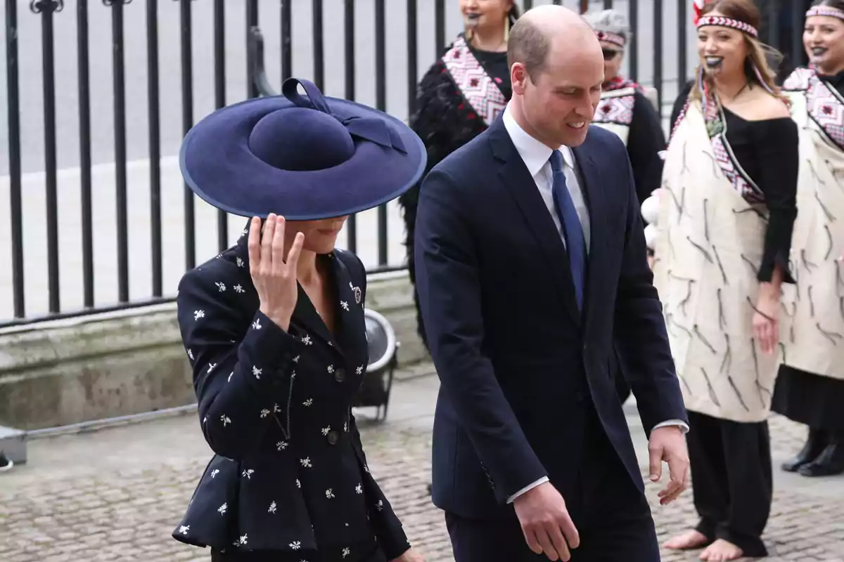 Kate Middleton, wearing a large blue hat, walks alongside Prince William in a dark suit while several people in traditional attire smile in the background.