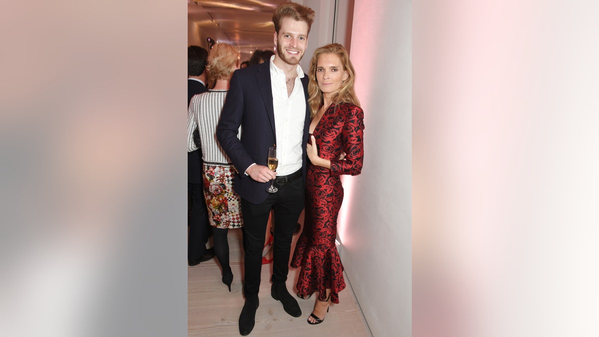 Louis Spencer in a black blazer, black pants and white shirt, holding a flute of champagne next to a woman wearing a dark red dress.