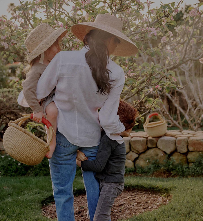 Woman wearing a wide-brimmed hat carrying children and a basket while fruit-picking in an orchard during daylight. Woman wearing a wide-brimmed hat carrying children and a basket while fruit-picking in an orchard during daylight.