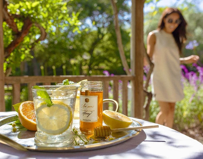 Close-up of a refreshing citrus drink and honey jar on a tray with a blurred woman in the background in a garden setting. Close-up of a refreshing citrus drink and honey jar on a tray with a blurred woman in the background in a garden setting.