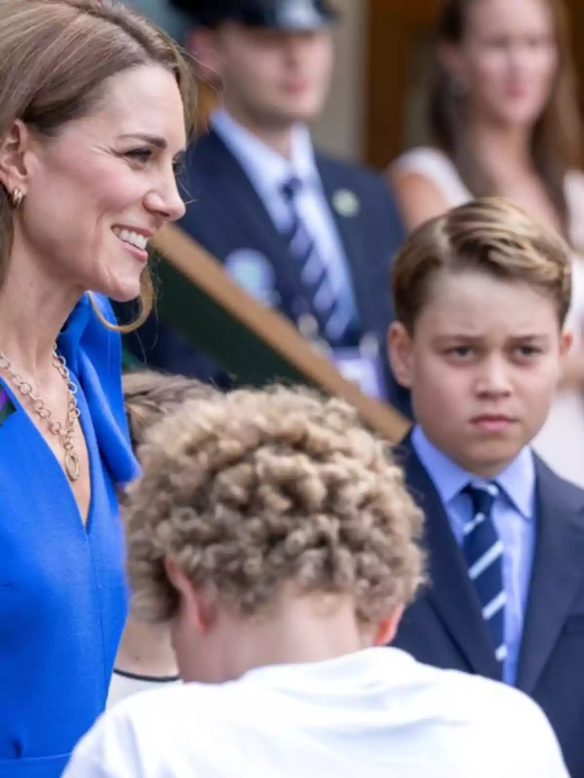 Kate Middleton, smiling in a blue dress, is accompanied by Prince George in a suit and tie and another curly-haired boy in the foreground, while several people watch in the background. Kate Middleton, smiling in a blue dress, is accompanied by Prince George in a suit and tie and another curly-haired boy in the foreground, while several people watch in the background.