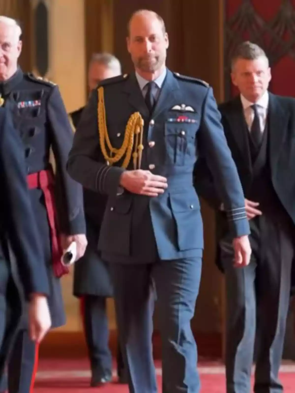 Prince William, dressed in a blue military uniform, walks down a carpeted hallway accompanied by other men in formal attire. Prince William, dressed in a blue military uniform, walks down a carpeted hallway accompanied by other men in formal attire.