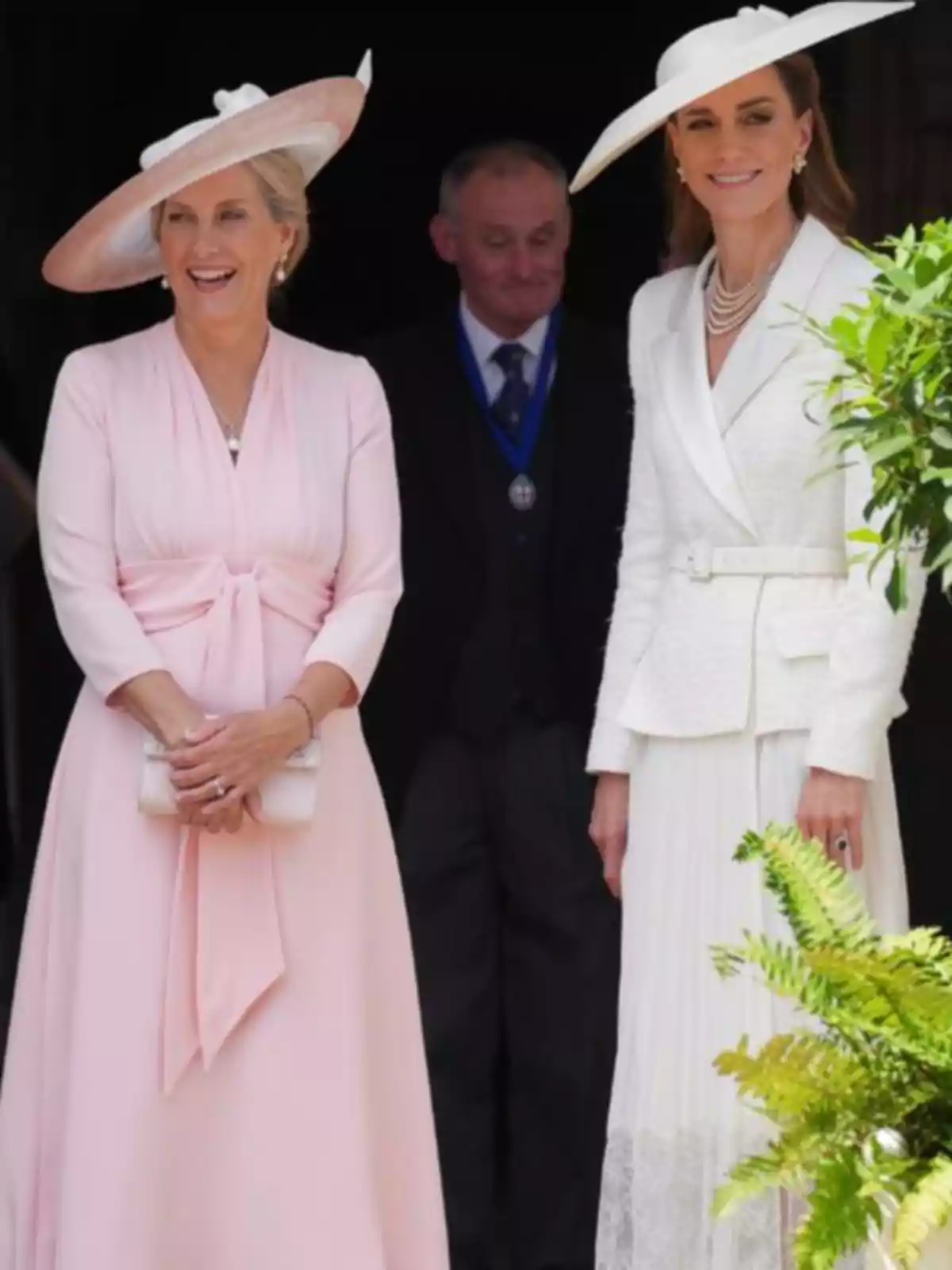Kate Middleton and another elegantly dressed woman wearing light-colored hats smile outdoors next to a man in a suit in the background.