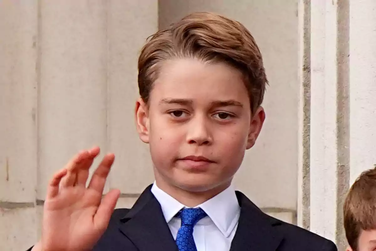 Prince George in a dark suit and blue tie raises his hand in front of a stone building.
