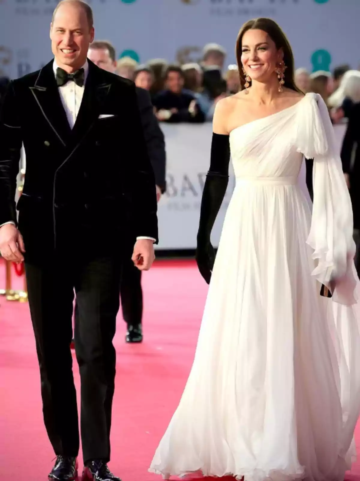 Elegantly dressed couple walking on a red carpet, he in a black tuxedo and she in a one-shoulder white dress with long black gloves
