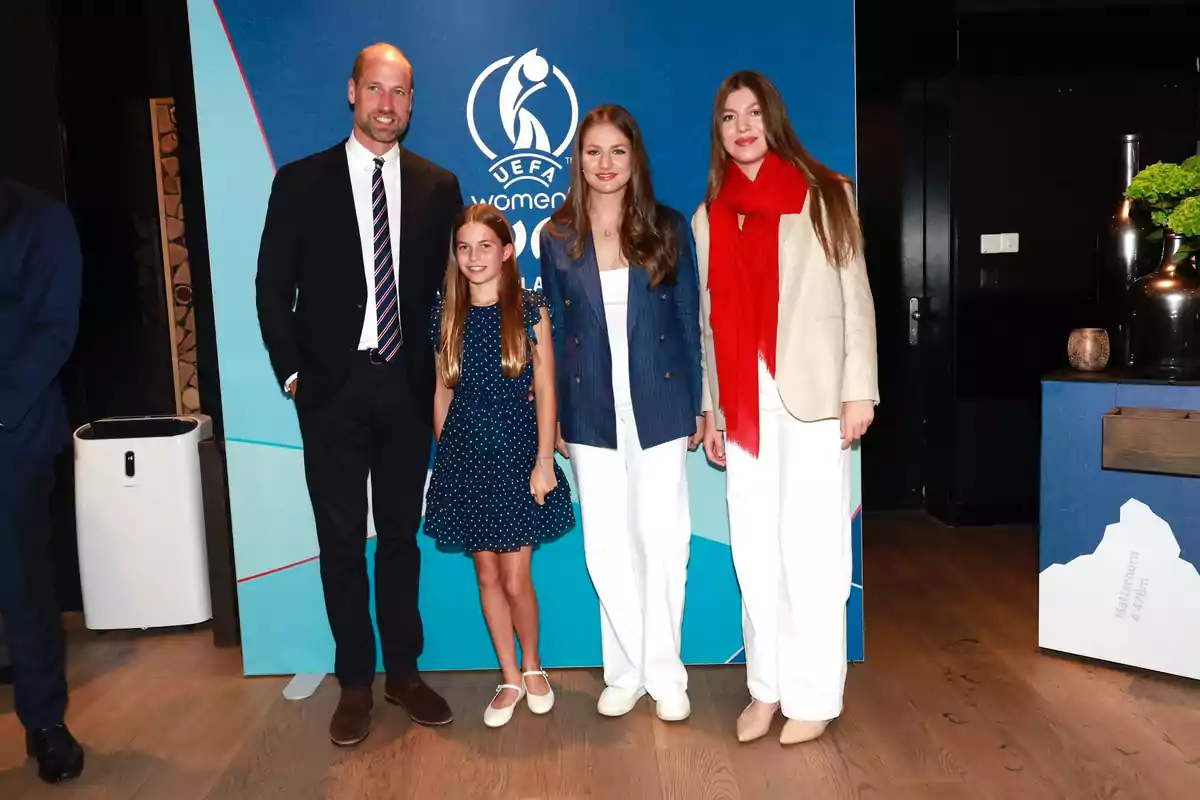 Leonor, Sofía, Prince William, and his daughter Charlotte pose smiling in front of a blue background with the UEFA Women’s Euro logo; two of them are wearing white pants and a red scarf, while a girl is wearing a blue dress with white polka dots.
