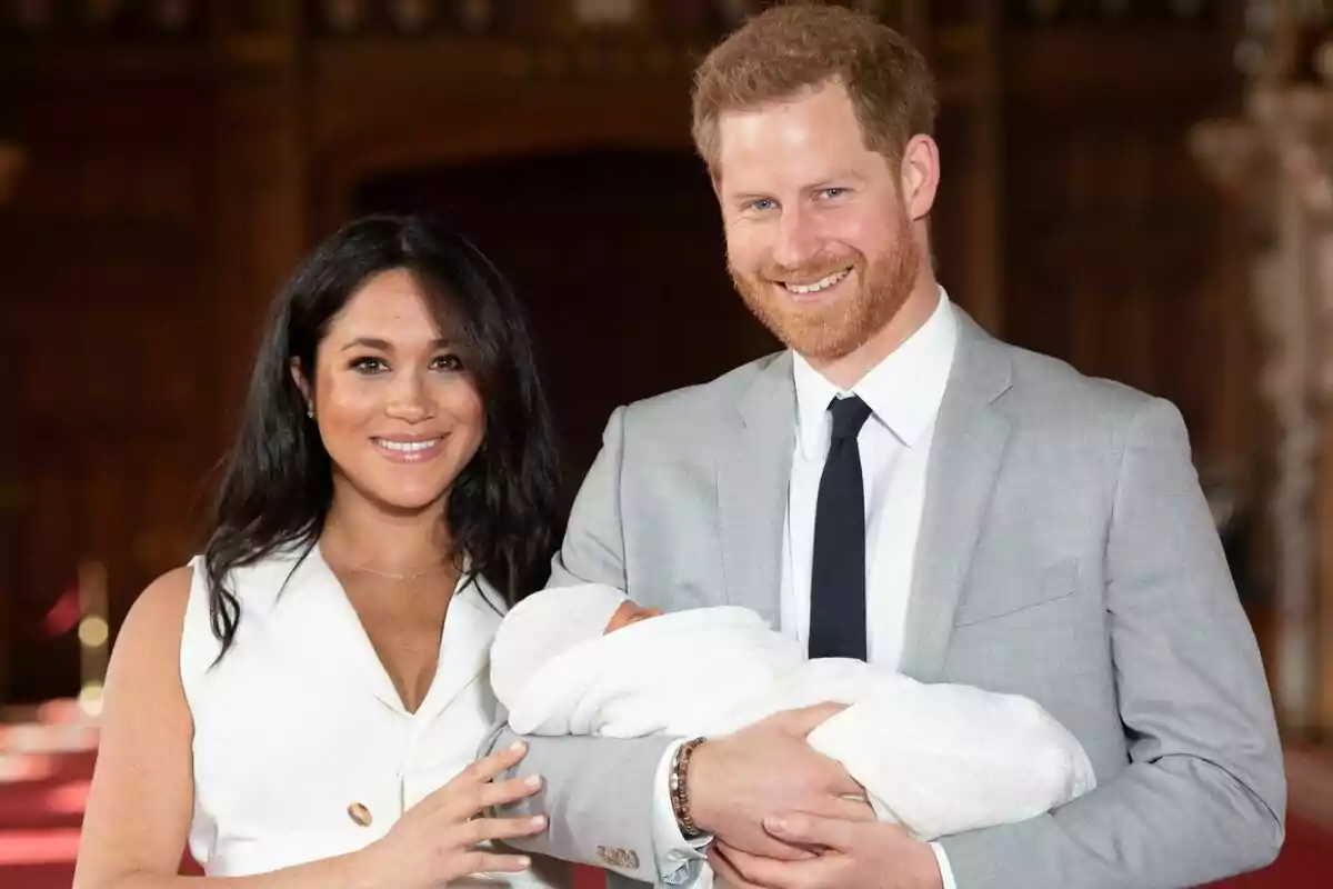 A smiling couple, Meghan Markle and Prince Harry, hold a baby wrapped in a white blanket.