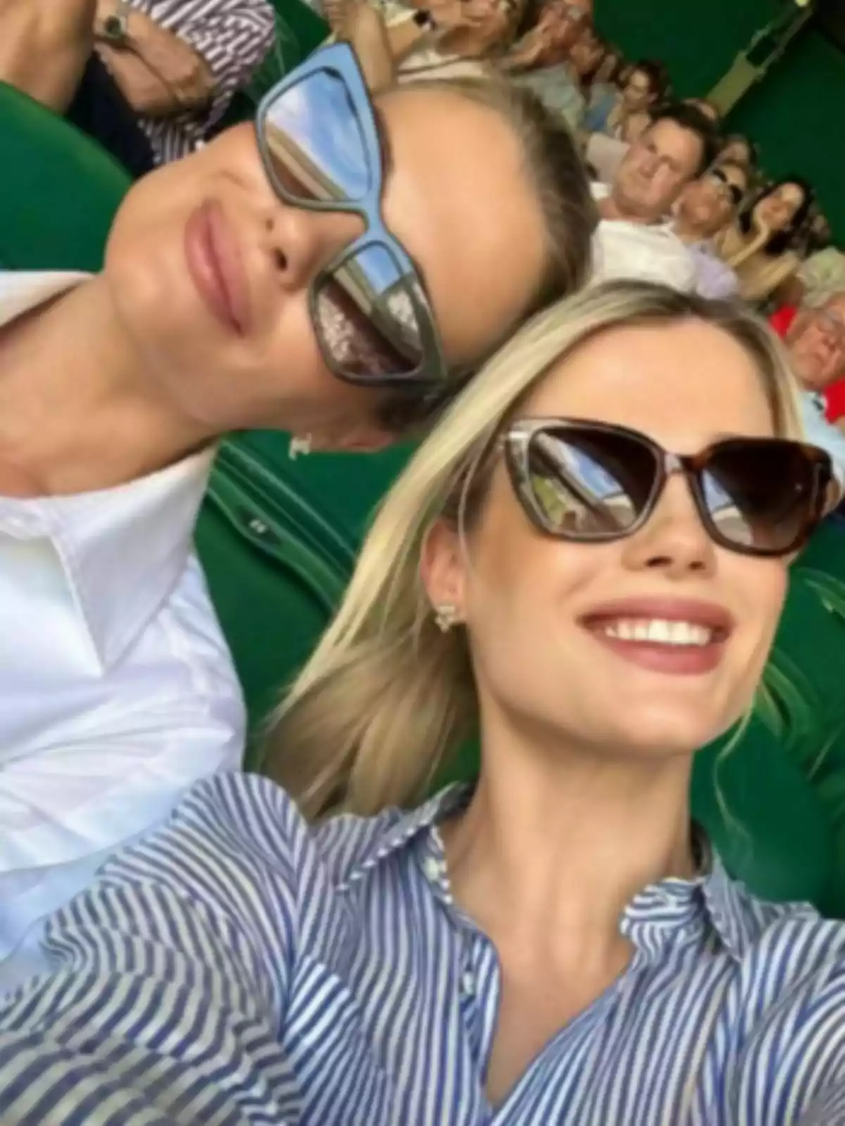 Two women smile wearing sunglasses, one of them is Amelia Spencer Mallett, as they take a selfie in a crowded stadium.