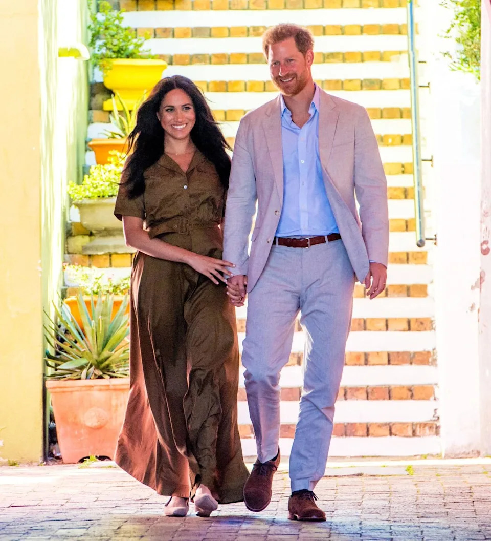 Meghan Markle and Prince Harry walk hand in hand through a colorful archway. Meghan wears a brown button-front dress and flats, while Harry wears a light gray suit with a blue shirt. Both are smiling as they step forward together.
