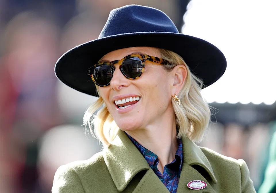 Zara Tindall wearing a blue fedora and sunglasses with a green coat and smiling