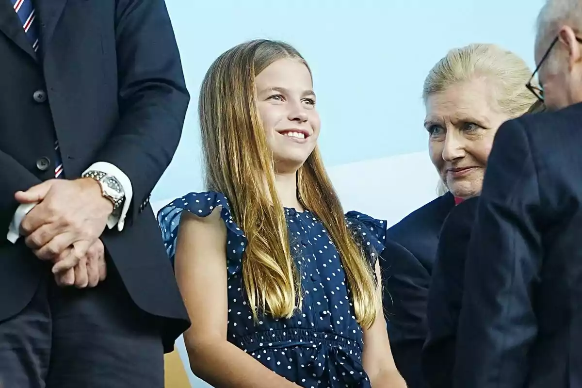 Smiling girl in a blue polka dot dress surrounded by adults at a formal event