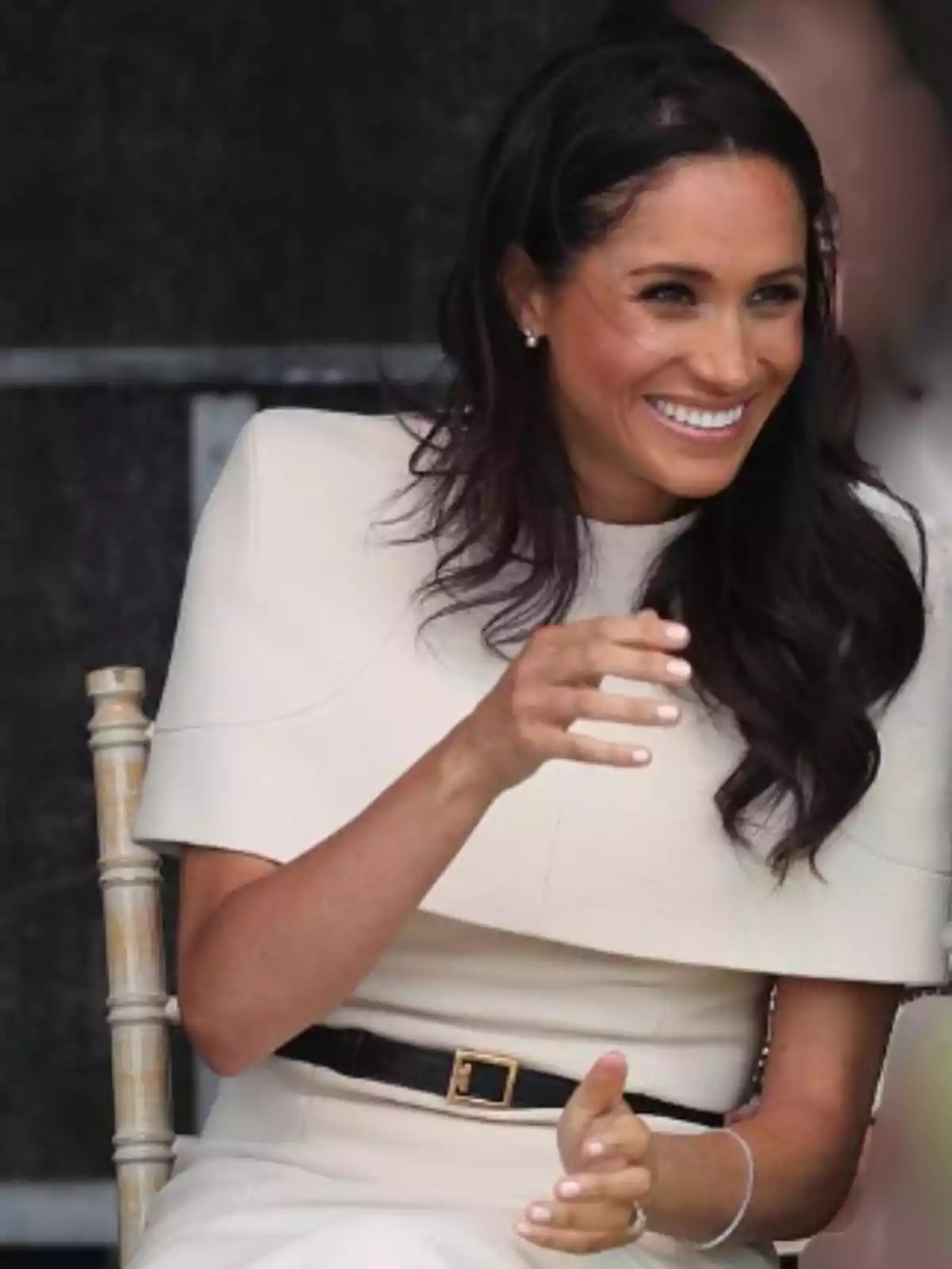 Dark-haired, long-haired woman sitting on a wooden chair, smiling and dressed in a cream-colored outfit