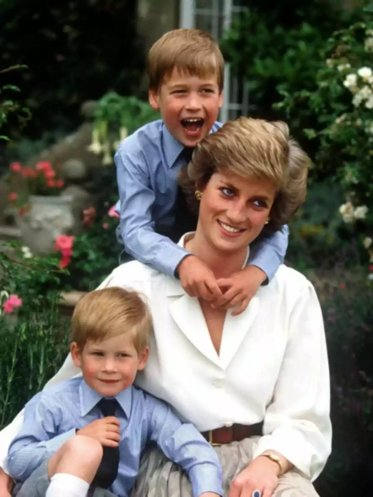 Smiling Lady Di sitting in a garden with Princes William and Harry hugging her, all dressed elegantly and surrounded by flowers.