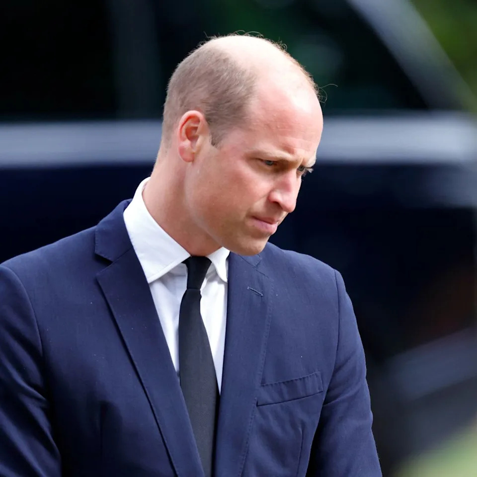 Prince William wears a navy suit and looks down at the ground, while wife Kate Middleton wears a black suit and has long brown golden hair.