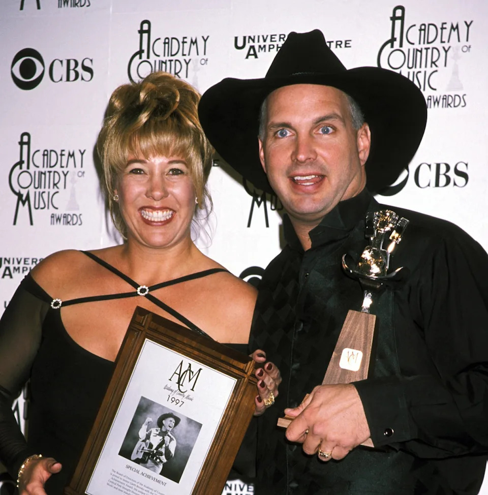 Two people smiling at the Academy of Country Music Awards. The person on the right wears a cowboy hat and holds a trophy