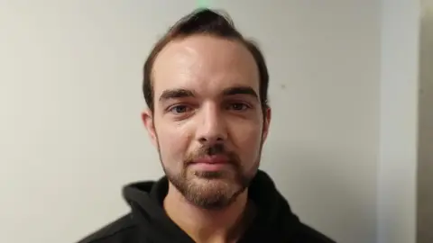 Micky Murray is standing by an off-white wall, he has a black hoodie on. Micky has short brown hair and a brown beard. He also has a piercing under his lower lip.
