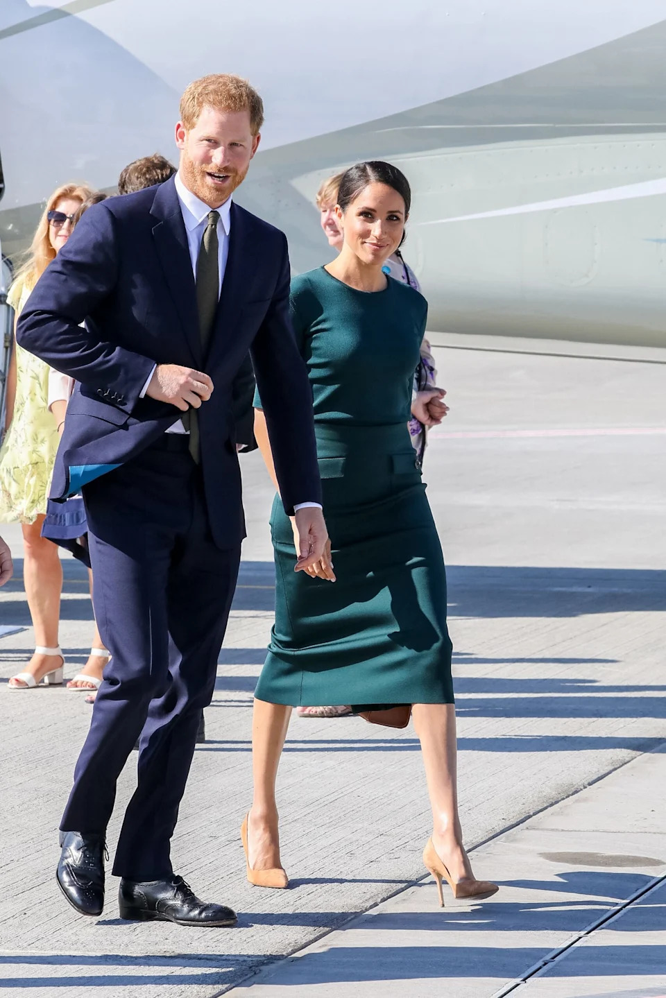 Prince Harry and Meghan Markle walk side by side on a sunny day at an airport. Harry wears a navy suit with a white shirt and tie while Meghan wears a fitted dark green dress with nude heels.
