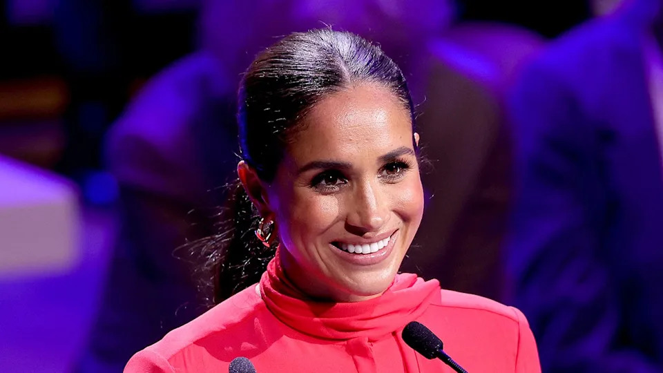A close-up of Meghan Markle smiling wearing a high collar red suit in front of a podium