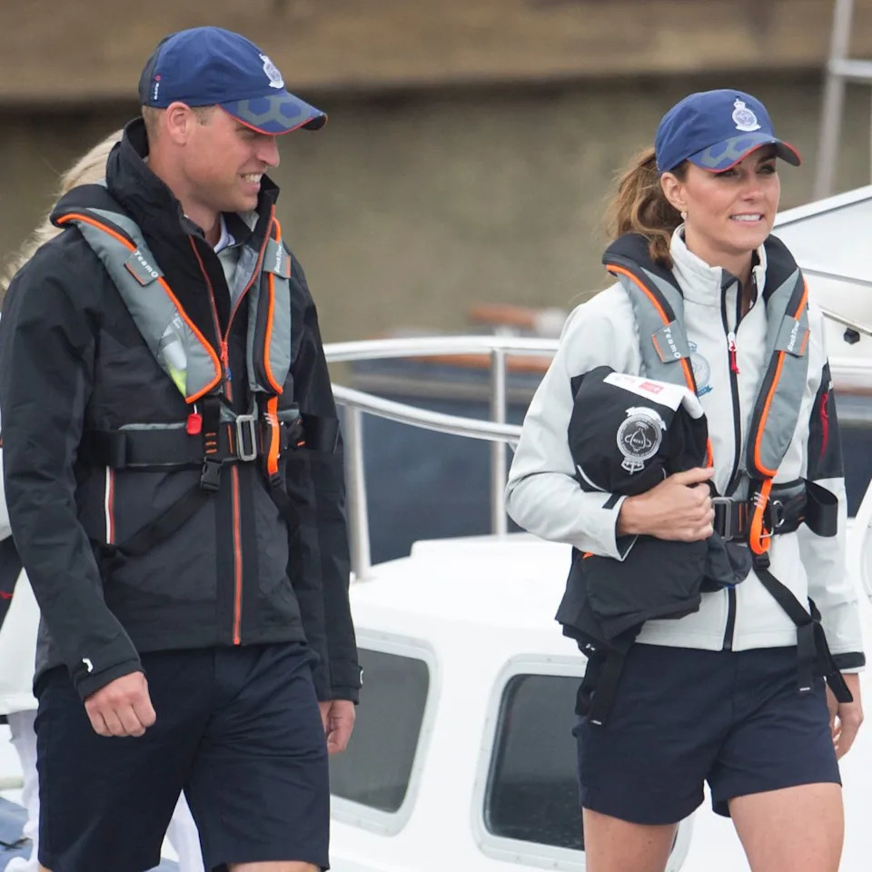  Prince William and Princess Kate wearing blue baseball caps and sailing gear and shorts walking on a dock. 