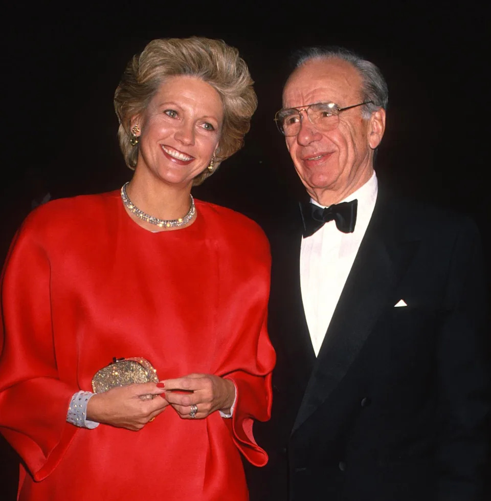 Two individuals on a red carpet event. The woman wears an elegant outfit, holding a clutch; the man is in a classic tuxedo, both smiling