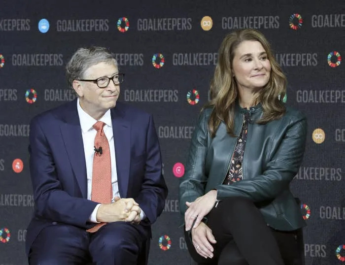 Two people are seated at a Goalkeepers event. One wears a suit with a tie, and the other wears a leather jacket and floral top