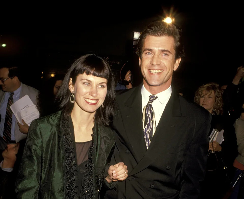 A smiling couple on a red carpet, the man in a suit with a patterned tie and the woman in a stylish embroidered jacket