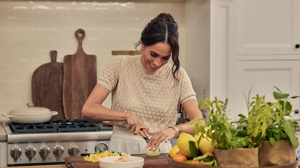 Meghan Markle wearing a beige sweater chopping vegetables.