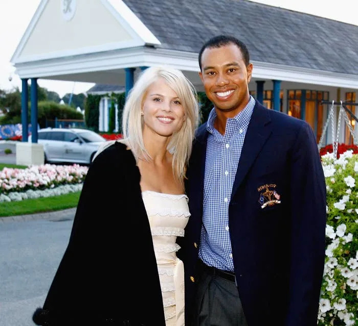 Two people are posing together outdoors; one wears a strapless dress with a shawl, and the other wears a blazer with a checked shirt