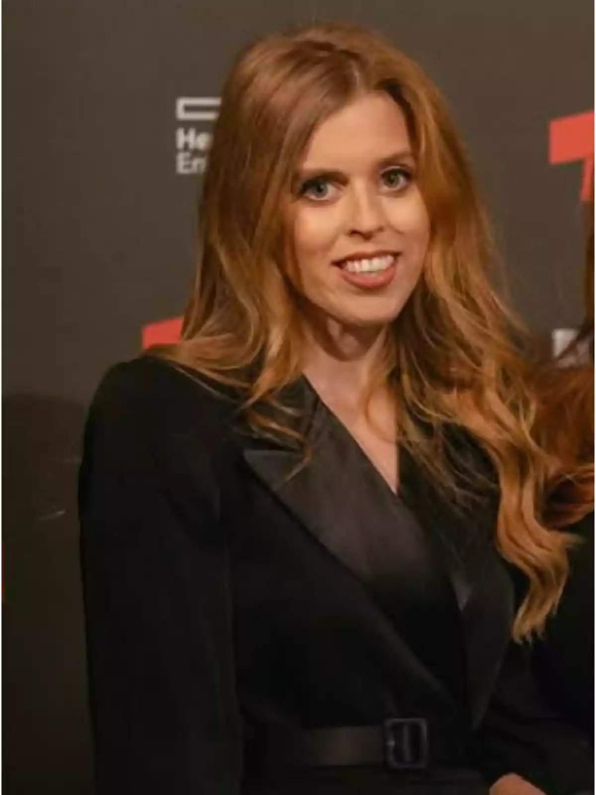 Long-haired blonde woman wearing a black jacket posing in front of a dark background at an event