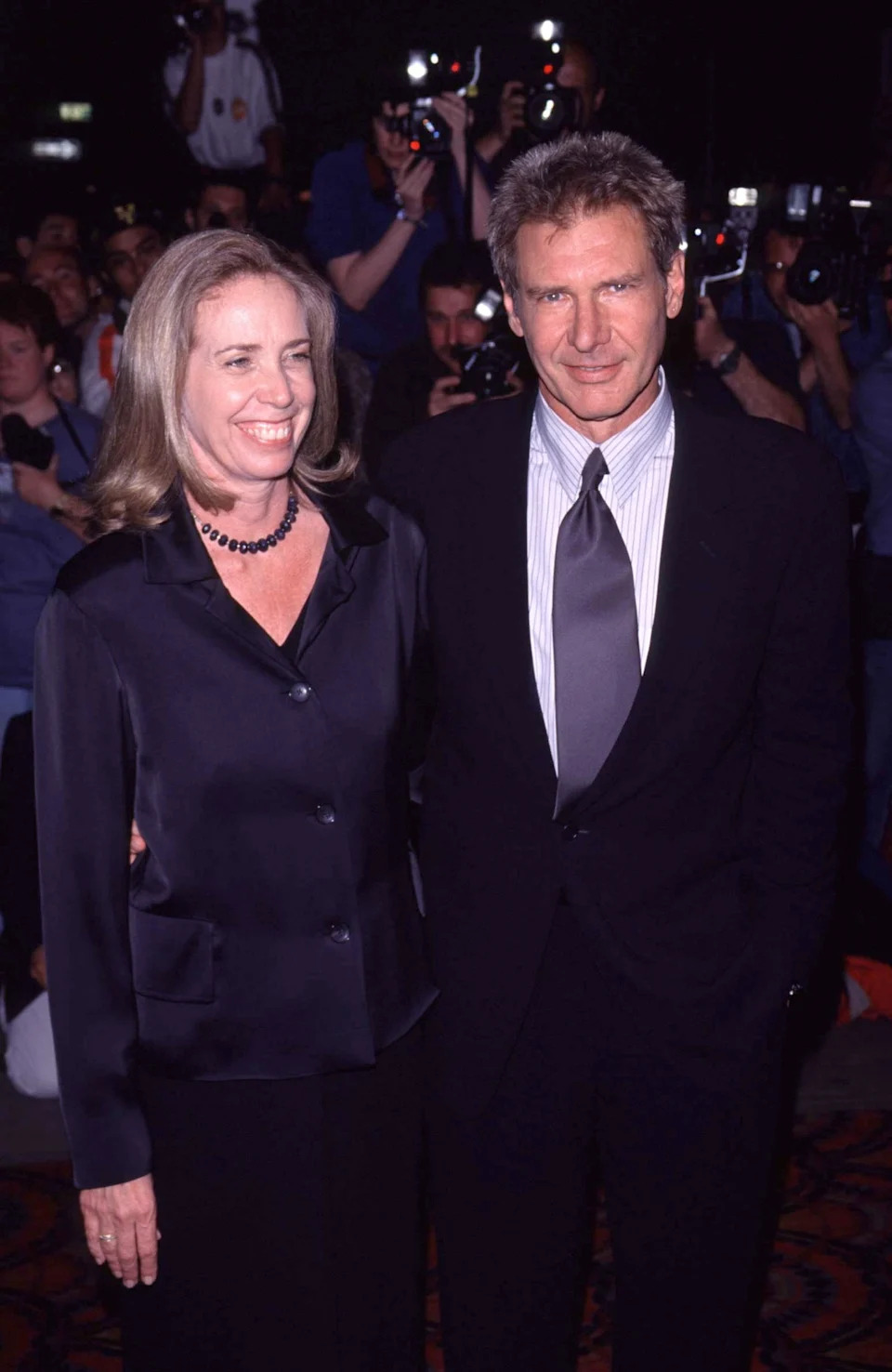 Two individuals pose at an event. The man wears a suit with a tie, and the woman is in a blouse with a necklace. Photographers are visible in the background