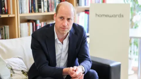 Getty Images A balding man in a blue suit is sat on a sofa. He has a straight face and is clasping his hands together in front of himself. He is mostly bald with very short brown hair on either side. 