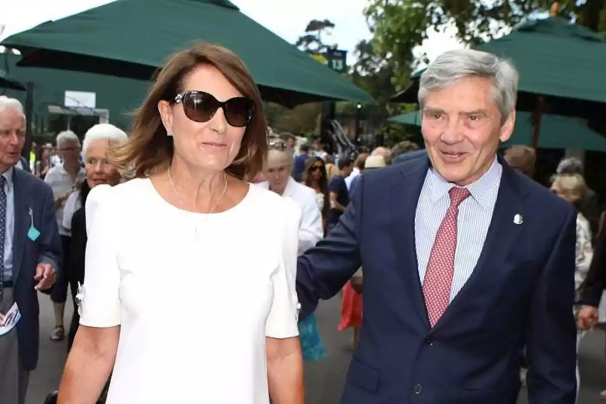A woman wearing sunglasses and a white dress walks next to a gray-haired man in a blue suit and red tie at an outdoor event surrounded by other people.