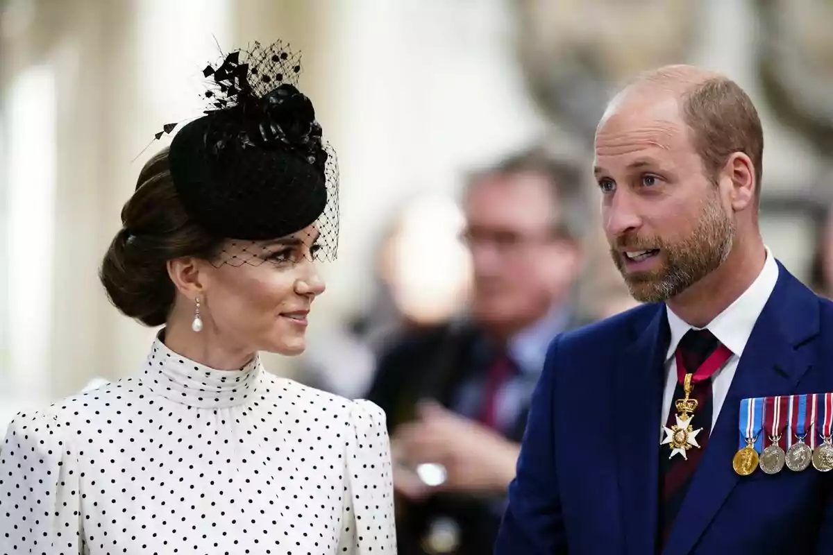 A woman in an elegant black hat and polka dot dress is talking to a man wearing a blue suit and several medals.
