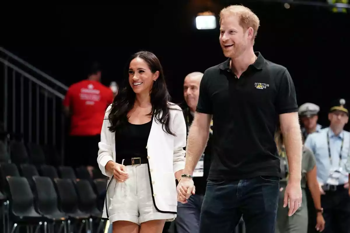 A smiling couple walks hand in hand at a sporting event with people in the background.