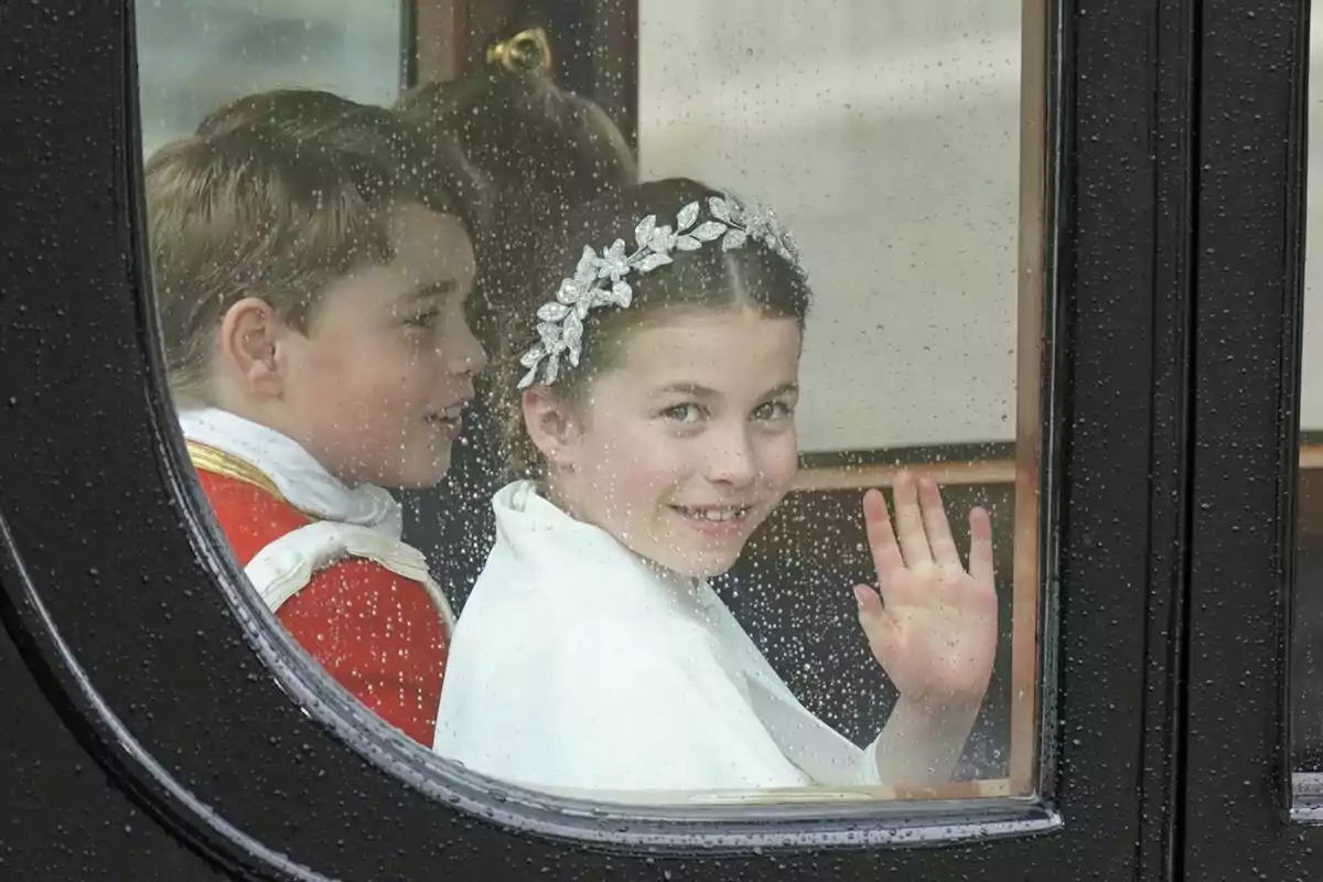Two smiling children inside a carriage wave through a window with raindrops.