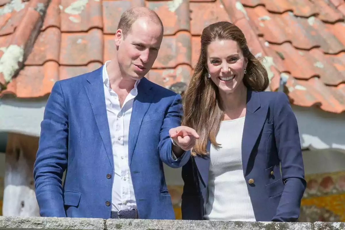 A smiling couple dressed elegantly in blue jackets posing in front of a tiled roof.