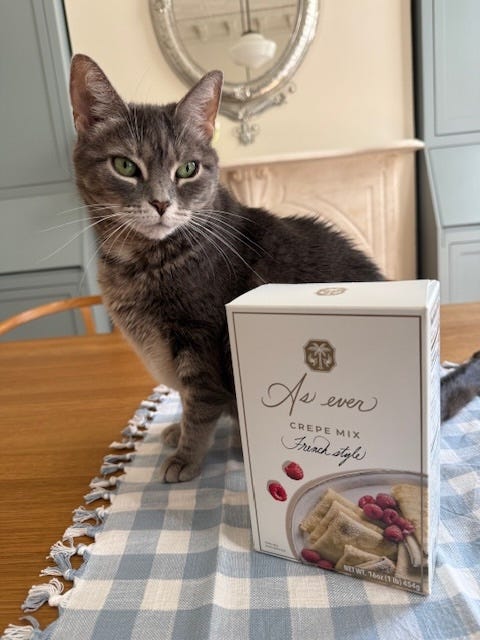 a cat positioned beside a box of crepe mix on a checkered tablecloth