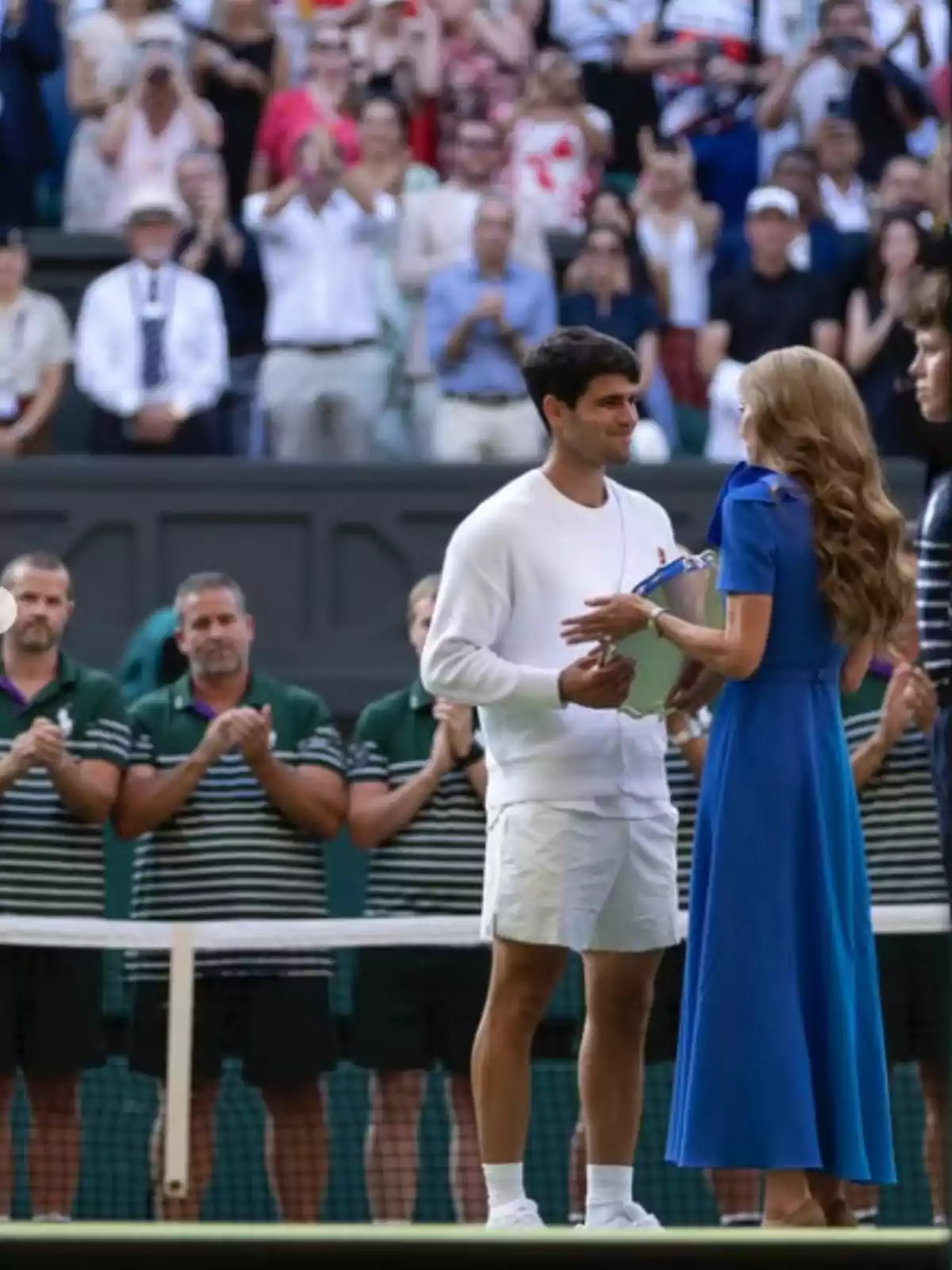 A tennis player receives a trophy from a woman dressed in blue on a tennis court while the crowd and the team applaud in the background.
