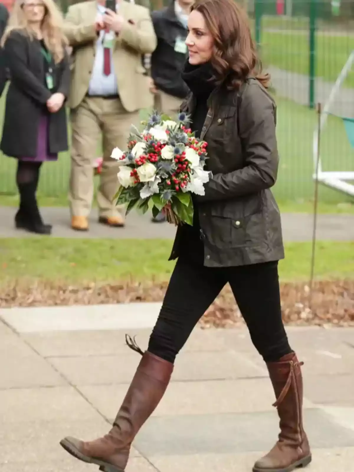 Woman walking outdoors with a bouquet of flowers in her hands, dressed in a dark jacket, black pants, and brown boots, while several people watch in the background.