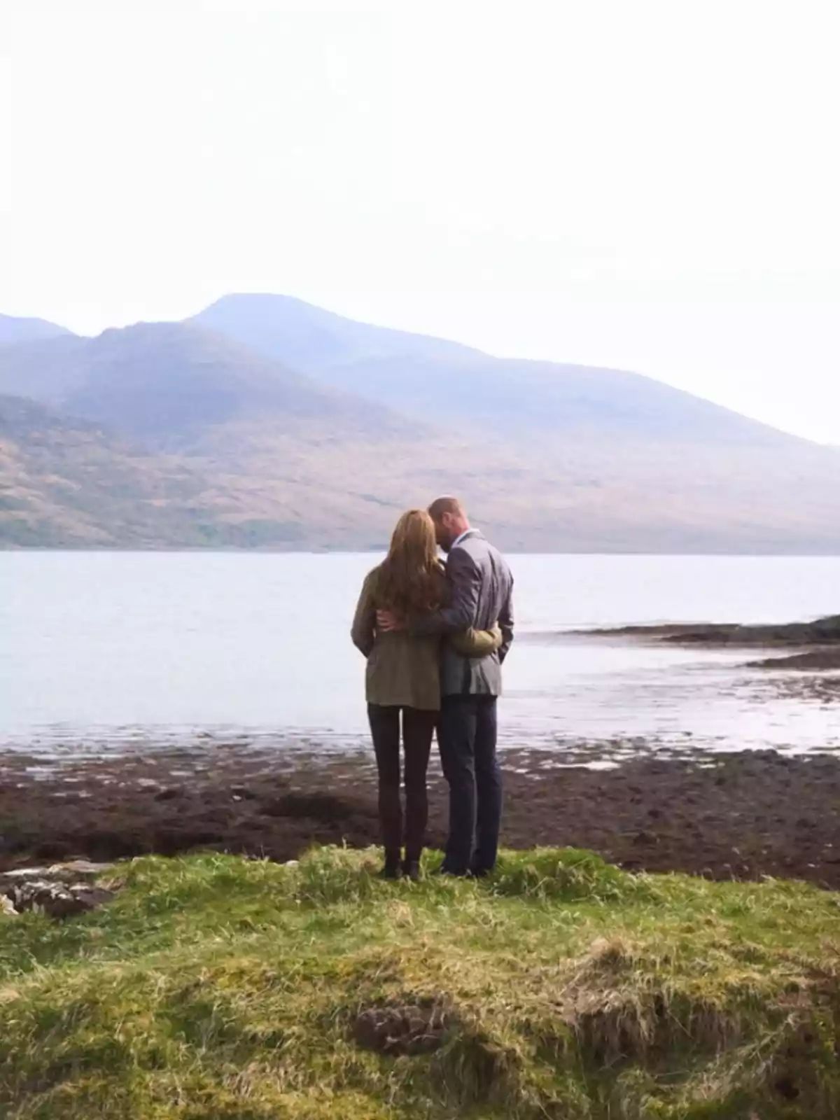 A couple embraced from behind looking at a lake with mountains in the background.