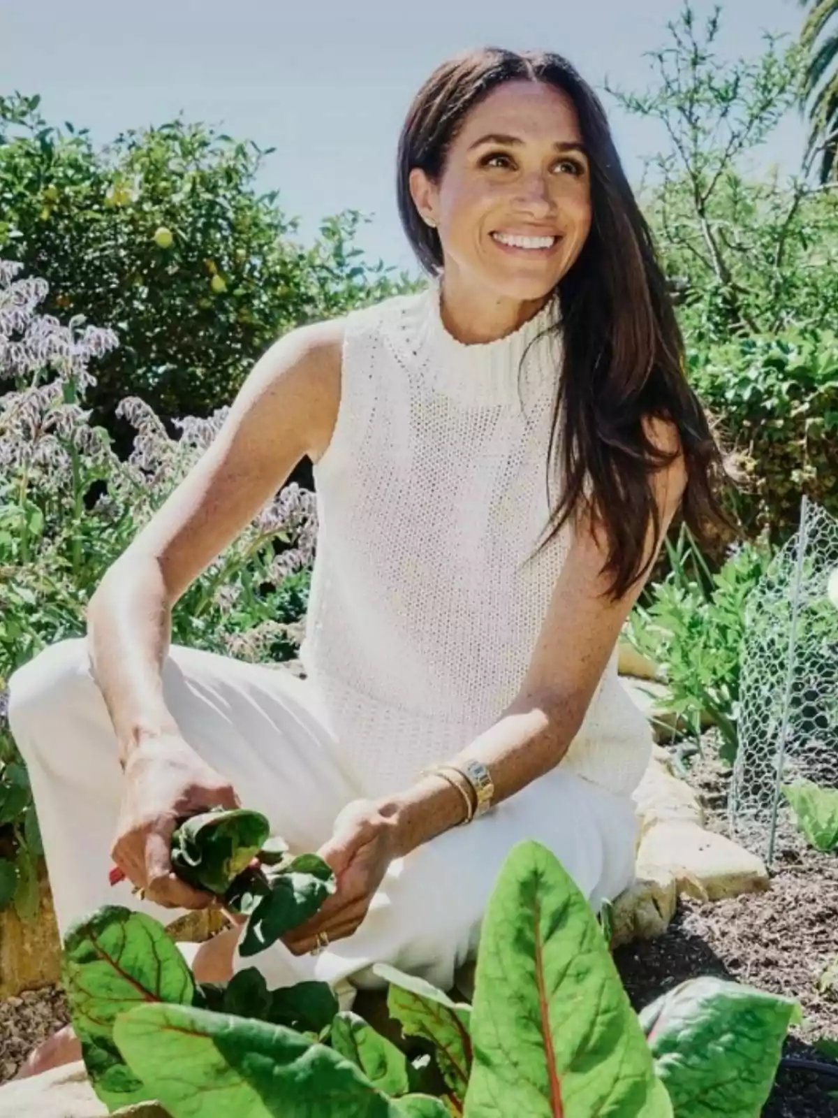 Smiling woman with long, loose hair dressed in white sitting in an orchard while harvesting green leaves, surrounded by plants and vegetation under a clear sky