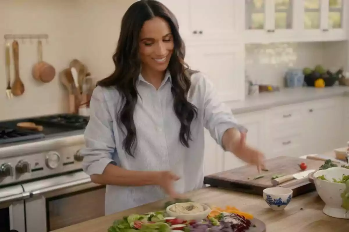 Meghan Markle smiles as she prepares a salad in a modern kitchen.