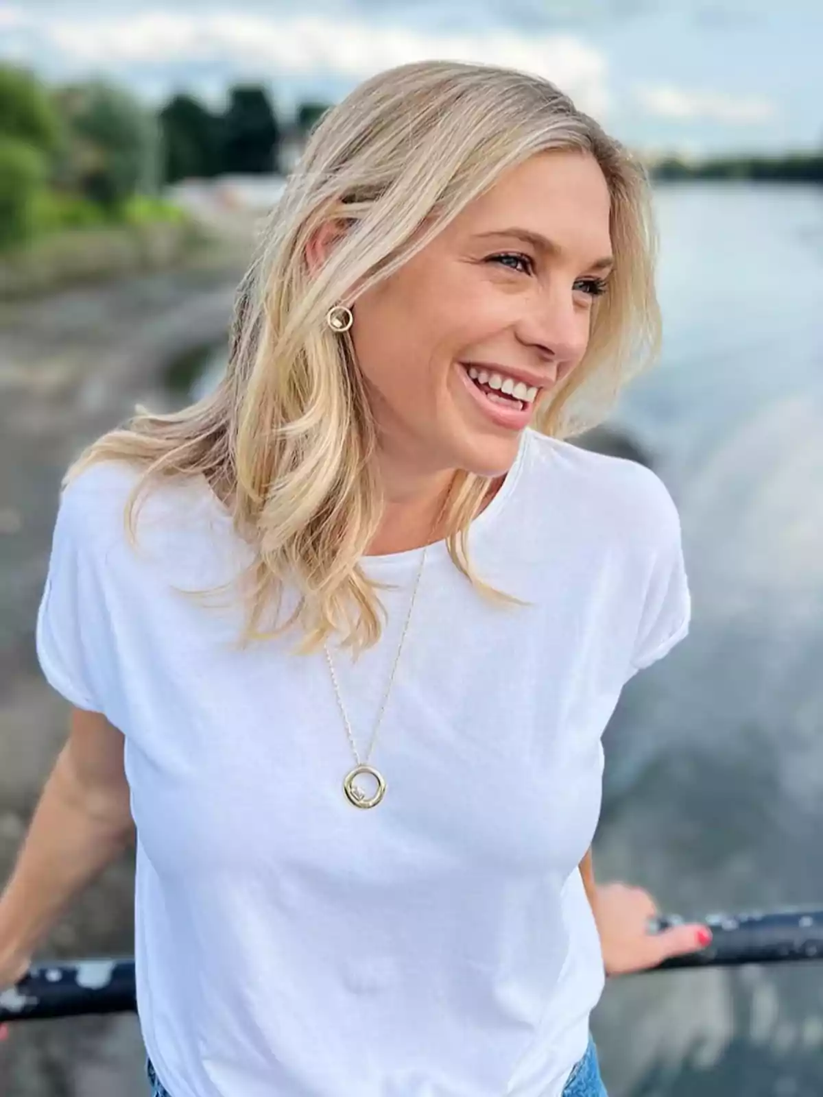 Chelsy Davy smiling in a white t-jersey and gold necklace, posing outdoors next to a body of water