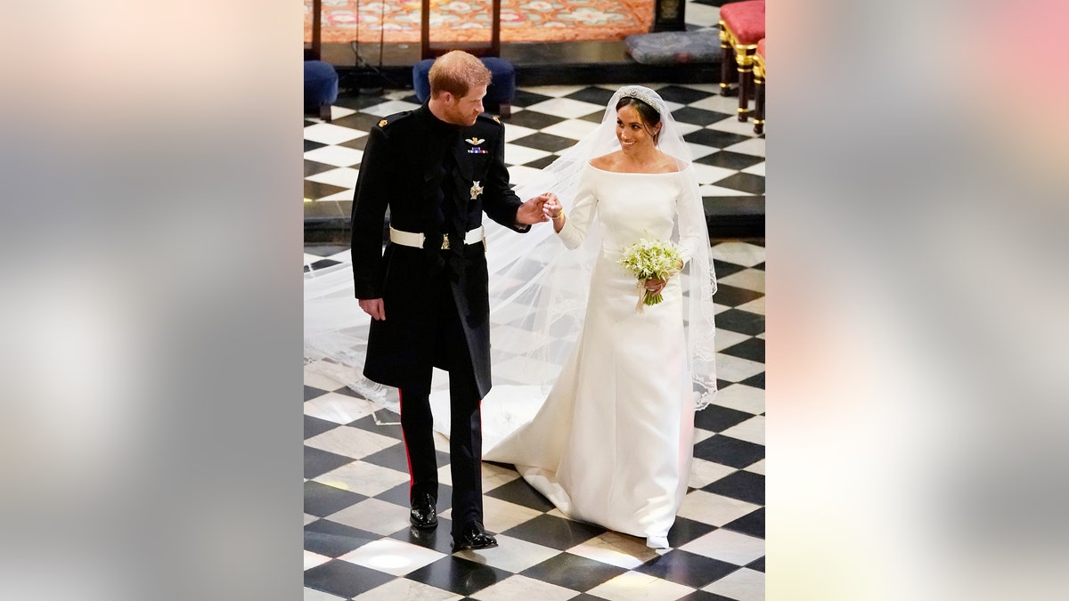 Prince Harry in a formal black suit holding Meghan Markle's hand on their wedding day.