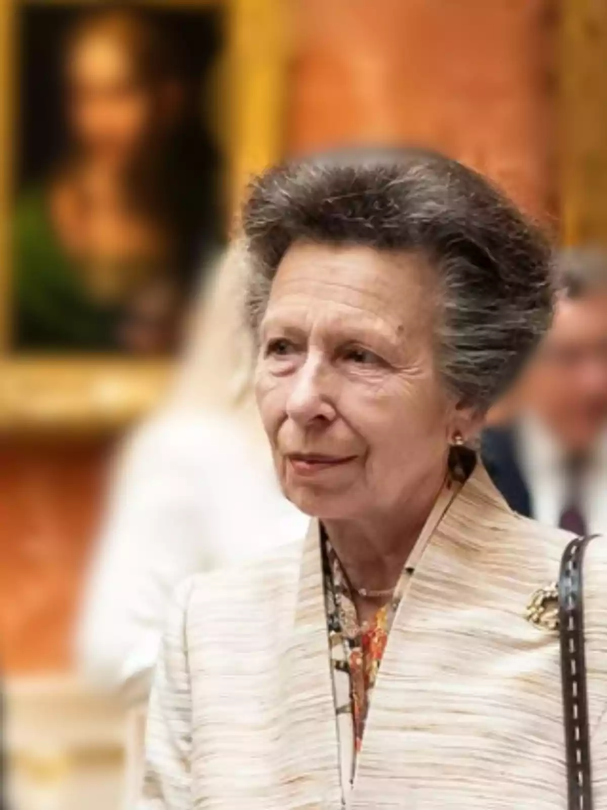 Elderly woman with gray hair styled in a voluminous updo, elegantly dressed with a gold brooch, in an art gallery with a blurred painting in the background
