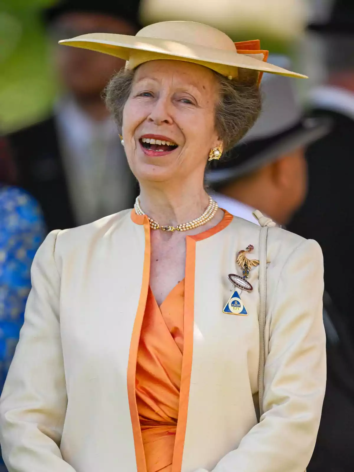 Princess Anne, smiling, wearing a beige hat and an elegant outfit in cream and orange tones, with a pearl necklace and brooches, at an outdoor event