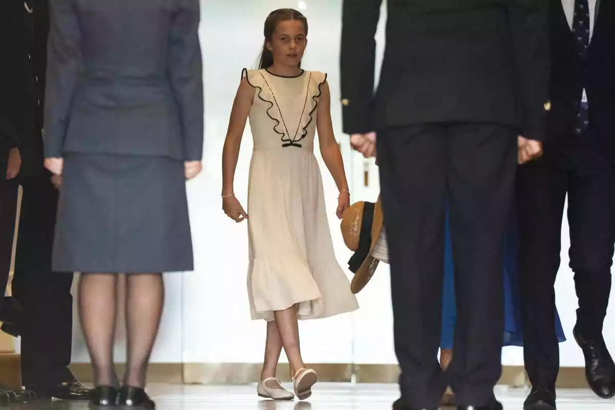Princess Charlotte in a light dress and white shoes walking down a hallway surrounded by adults dressed formally.