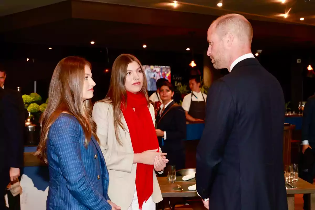 Leonor and Sofía talk with Prince William in an elegant setting with people in the background.