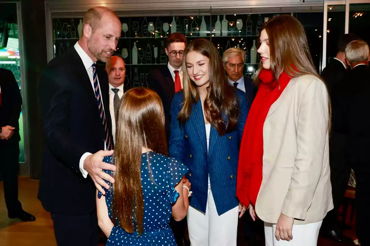 A man in a suit greets a girl while two young people smile and watch the scene in a formal setting with several people in the background.