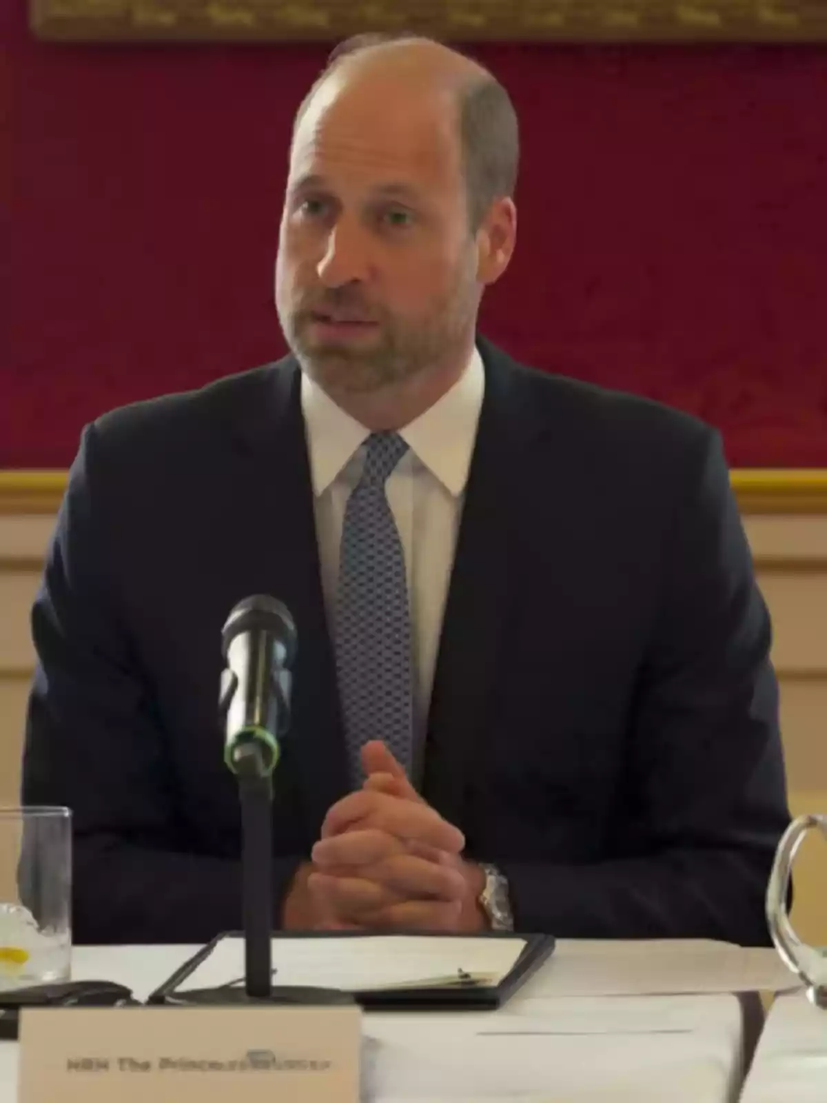 Man in a suit and blue tie speaking in front of a microphone at a conference table